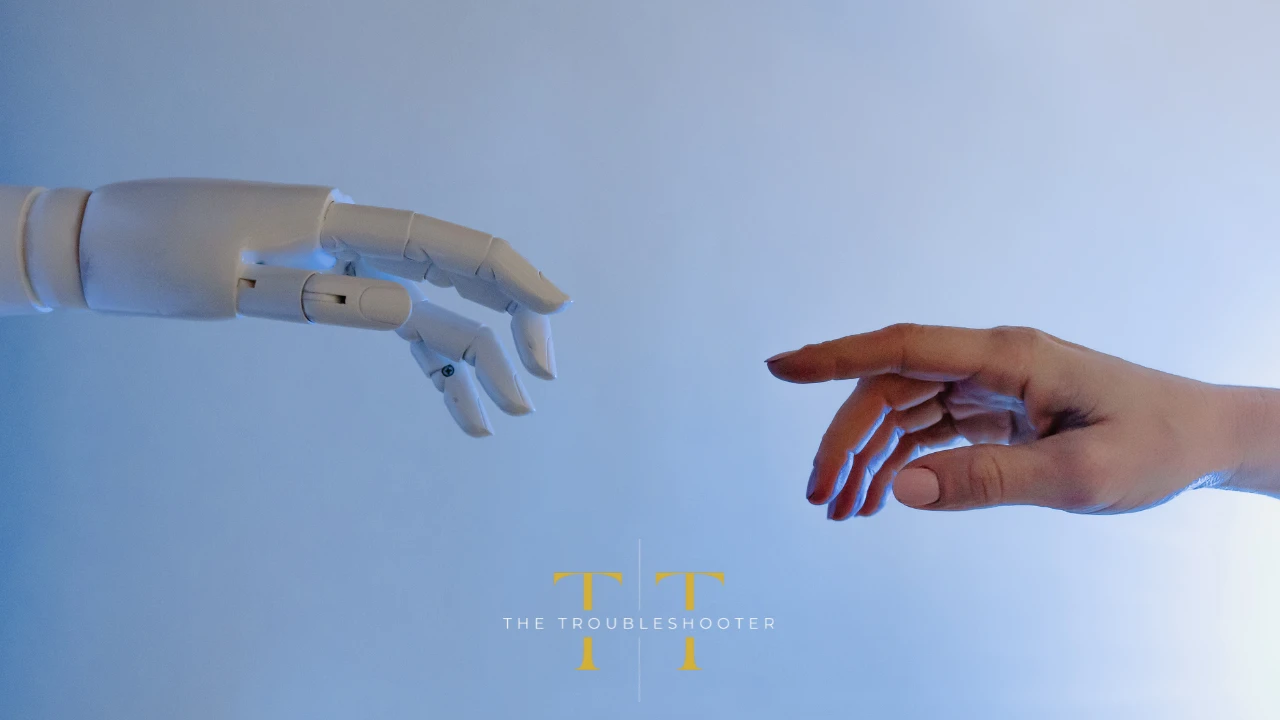 A robot hand reaches out to a human hand. The Troubleshooter logo in the foreground.