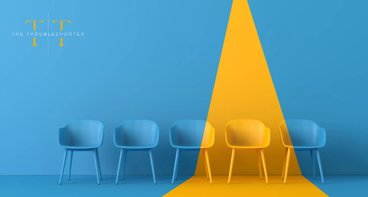 Five office chairs lined up in a row on a blue and yellow background. The Troubleshooter logo is top left.